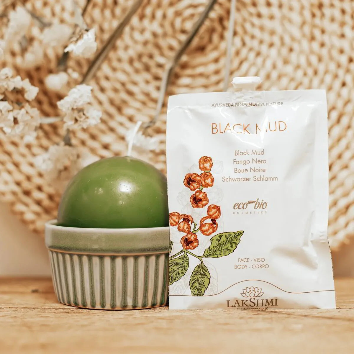 Lakshmi Black Mud sachet resting against a green ceramic vase on a woven mat